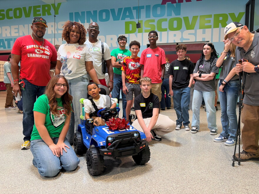 Several people pose for a picture next to a young boy. The young child is seated in a blue battery-powered ride toy car with a bow on top.