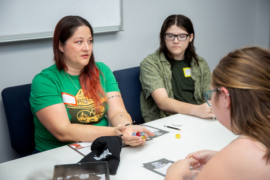 A woman with long, red hair and wearing a green t-shirt holds a bunch of brightly colored dice in her hand. 
