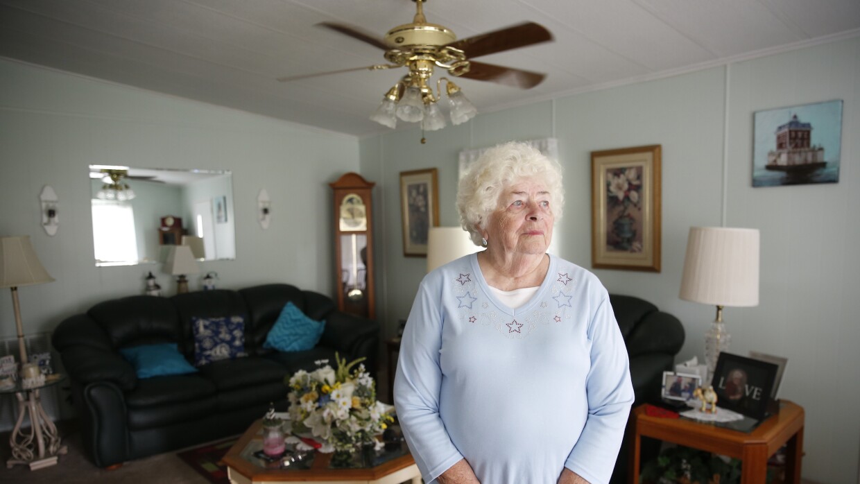 An elderly woman stands in front of her living room with a couch and other decorations in the background. 