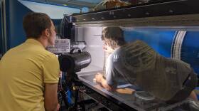 Two graduate student researchers Rhett Parry (left) and Mike Wayman (right) calibrate the wind tunnel, a large plastic and metal structure within a lab with a tree trunk in the middle