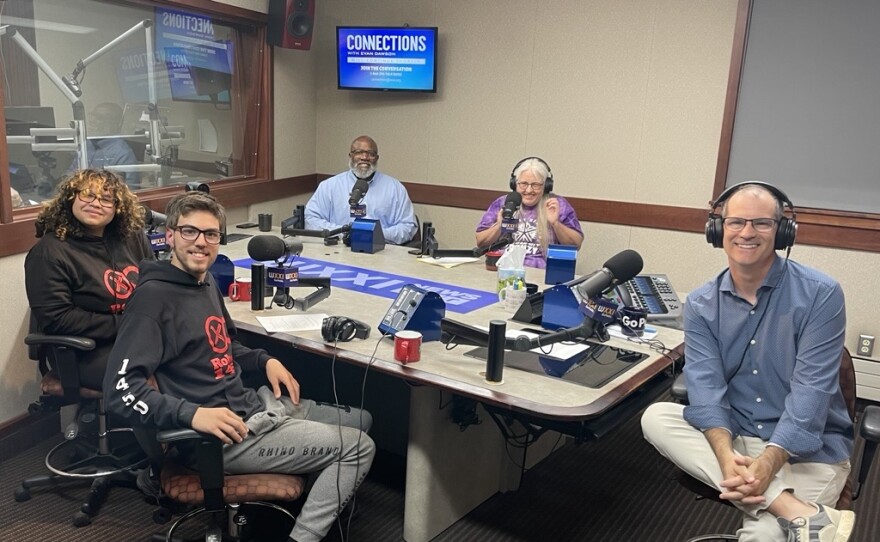 Five smiling people sit around a table in a radio talk studio: A young woman front left has shoulder-length curly black and blonde hair and is wearing glasses and a black hoodie with a red graphic; a young man front center has short brown hair and is wearing glasses, a black hoodie with a red graphic, gray sweatpants, and black shoes; a man back left is bald and has a white beard and is wearing a light blue button-down shirt; a woman back center has long white hair and is wearing glasses, a purple t-shirt with a white Rochester Flower City logo; a man at right has very short dark hair and is wearing headphones, a blue button-down shirt, white linen pants, and sneakers