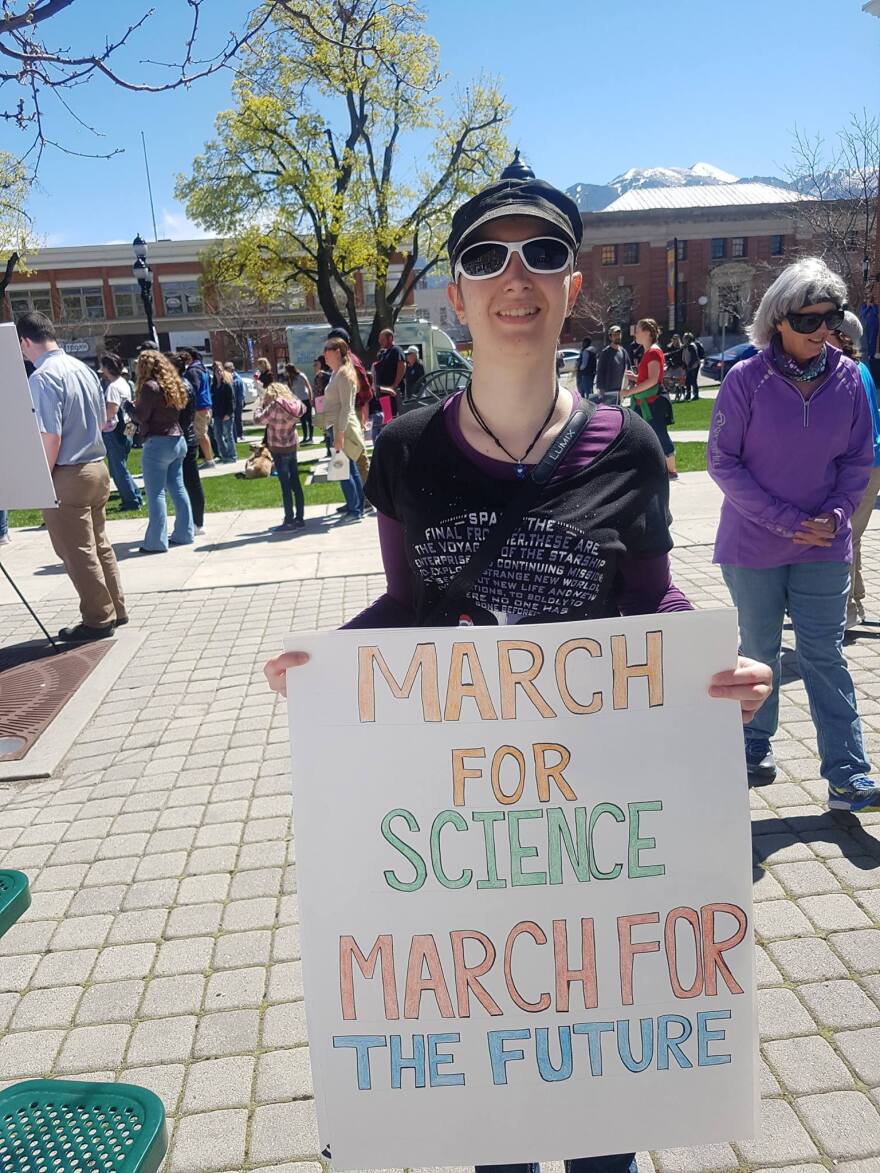 Background: blue sky, green tree and a mountain peak. Mid-ground: Bustling people on a cobblestone plaza. Foreground: Person holding a sign that says "March For Science. March For The Future"