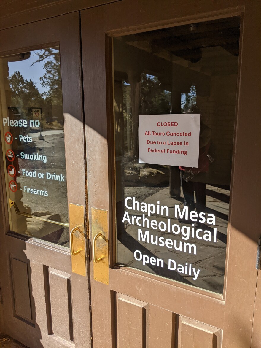 A brown wooden door frame with a glass top half reads Chapin Mesa Archeological Museum - Open Daily. A sign taped inside reads CLOSED All Tours Canceled Due to a Lapse in Federal Funding. The opposite door reads Please no - Pets - Smoking - Food or Drink - Firearms