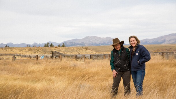 Two women stand in a dry field posing together for a photo in front of mountains in the distance.
