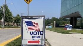  A sandwich board with an image of a waving American flag and the words "vote here" in block lettering directs people to a polling place in Kansas City. In the background campaign signs line the sidewalk. 