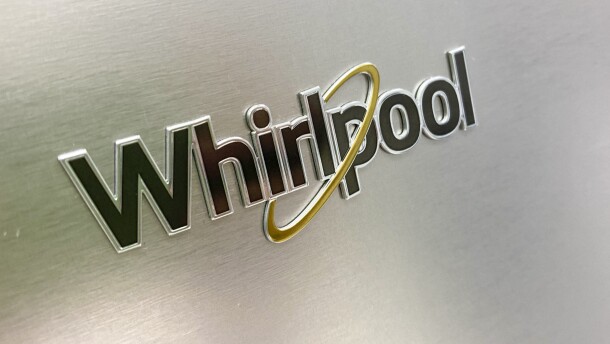 The Whirlpool logo is seen on a clean, bright gray metal surface. Whirlpool is spelled out with steel gray trimmed black text, with a yellow oval looping around the middle of the text. 
