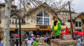 A large crowd stands in front of a building while a child in a dinosaur costume plays outside.