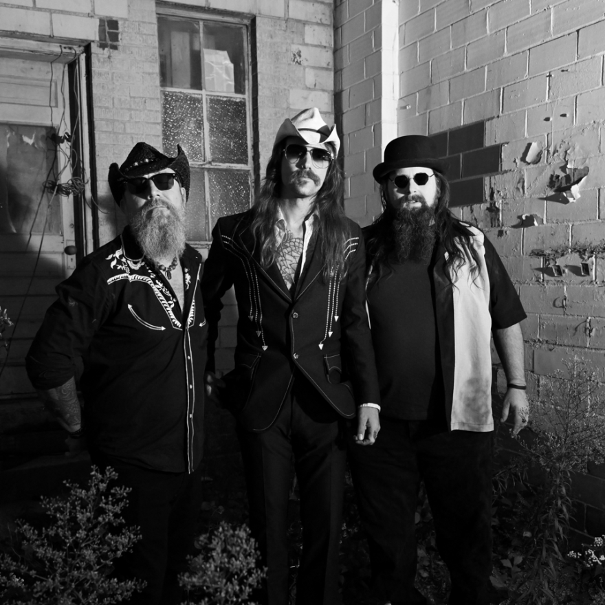A posed black and white image of the three members of The Other Brothers, wearing sunglasses, with a decrepit building right behind them. 