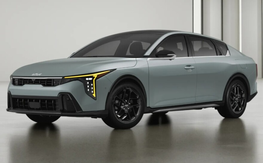 A studio photograph of a sleek, light grey or silver-blue modern sedan, positioned at a three-quarter angle facing left against a minimalist white background. The vehicle features striking, amber-colored geometric LED daytime running lights, a bold dark front grille, a black roofline, and intricate black multi-spoke alloy wheels.
