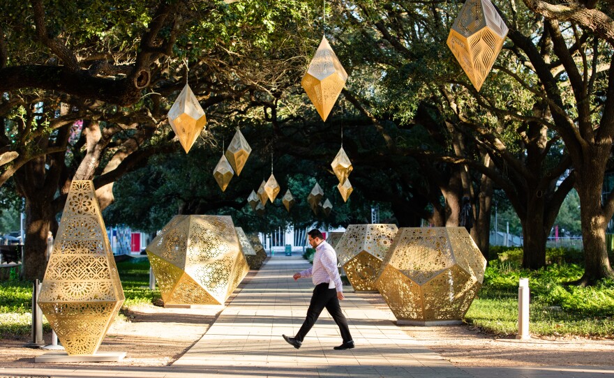 A man in casual business attire walks in front of several geometric art pieces in a tree-filled park during the day.