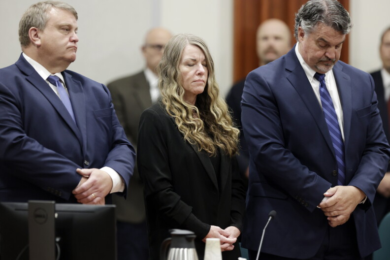 A woman with long blonde hair wearing all black standing between two men. They all have their hands crossed in front of them.