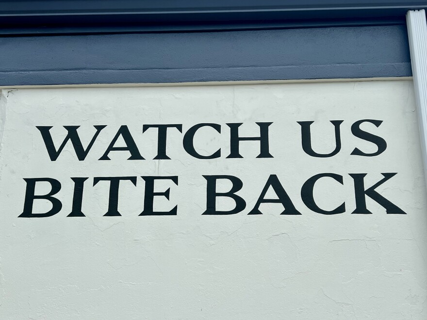 Black capitalized serif letters on a white wall that read, "Watch us bite back."