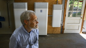 Despite his solar-charged batteries not yet being fully operational, Paul Aho (foreground) remains enthusiastic about the technology’s potential and the Energy Storage Solutions program that helped finance them. But, Aho says “Anyone going into this should be cautioned that it’s going to be a frustratingly long period of time before the batteries are actually working.” Aho made a deposit in January for two battery modules (background) through the state’s Energy Storage Solutions program.