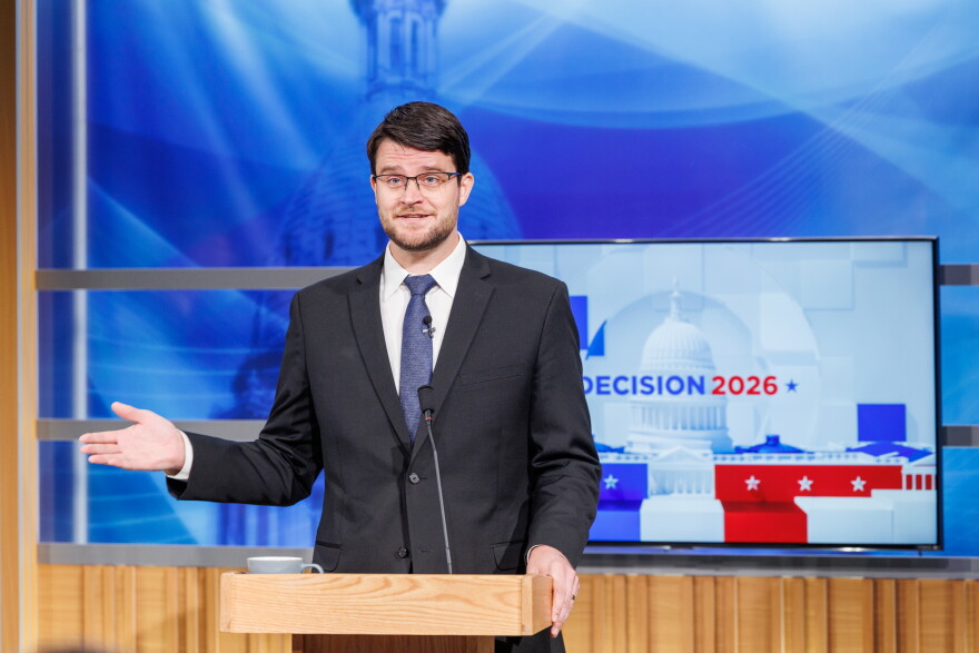 A man in a suit speaks at a podium in front of a TV that reads "Decision 2026"