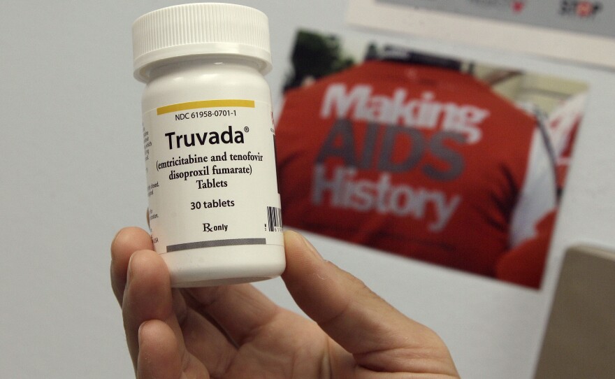 A hand holds a bottle of the drug Truvada. A sign says "Make AIDS History" in the background. 