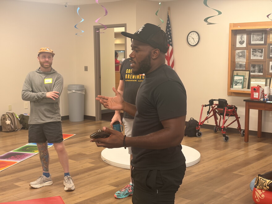 A man dressed in black and a black ball cap talks while two men look at him.
