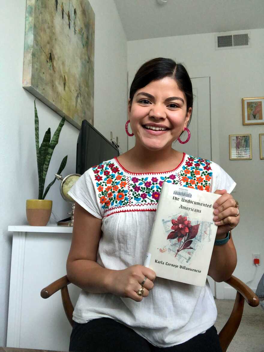 Reporter Alejandra Martinez sits in her home in a wooden chair holding her book recommendation: The Undocumented Americans by Karla Cornejo Villavicencio