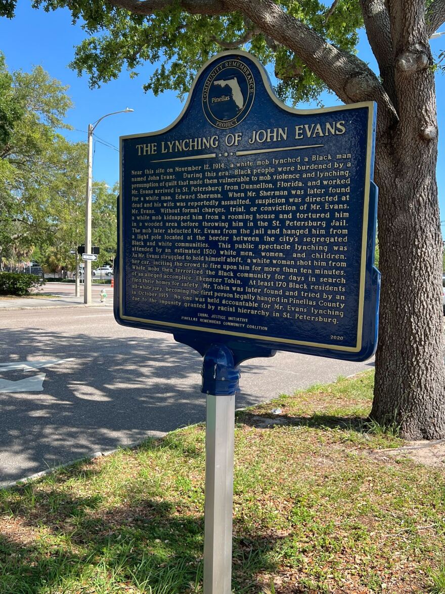 A dark blue sign with gold text that reads: "The Lynching of John Evans. Near this site on November 12, 1914, a white mob lynched a Black man named John Evans. During this era, Black people were burdened by a presumption of guilt that made them vulnerable to mob violence and lynching. Mr. Evans arrived in St. Petersburg from Dunnellon, Florida and worked for a white man, Edward Sherman. When Mr. Sherman was later found dead and his wife was reportedly assaulted, suspicion was directed at Mr. Evans. Without formal charges, trial, or conviction of Mr. Evans, a white mob kidnapped him from a rooming house and tortured him in a wooded area before throwing him in the St. Petersburg Jail. The mob later abducted Mr. Evans from the jail and hanged him from a light pole located at the border between the city’s segregated Black and white communities. This public spectacle lynching was attended by an estimated 1500 white men, women, and children. As Mr. Evans struggled to hold himself aloft, a white woman shot him from her car, inciting the crowd to fire upon him for more than ten minutes. White mobs then terrorized the Black community for days in search of an alleged accomplice, Ebenezer Tobin. At least 170 Black residents fled their homes for safety. Mr. Tobin was later found and tried by an all-white jury, becoming the first person legally hanged in Pinellas County in October 1915. No one was held accountable for Mr. Evans’ lynching due to the impunity granted by racial hierarchy in St. Petersburg."