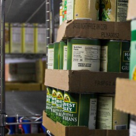 Cans of green beans and corn are stacked on metal shelves. 