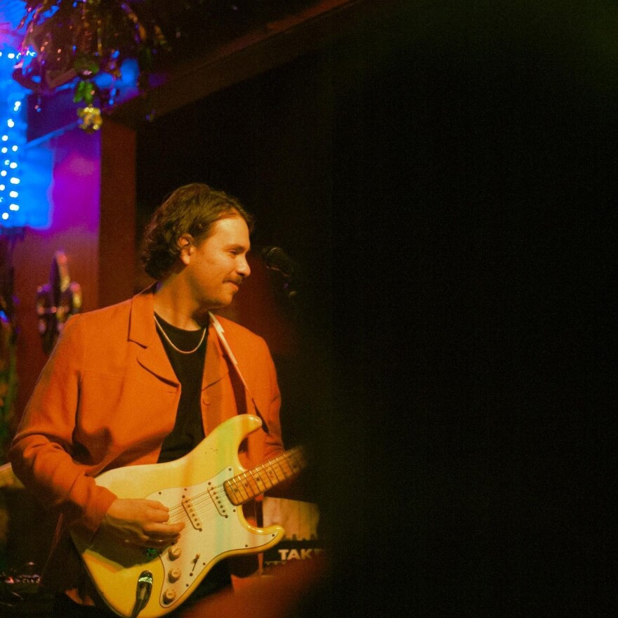 A musician in an orange jacket plays a white electric guitar on a dimly lit stage.