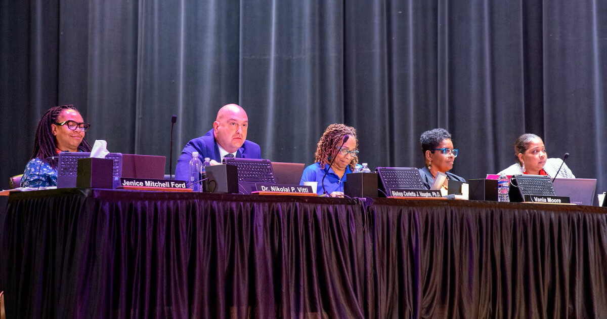 Five adults in suits sit in a line on an auditorium stage at a table with a black curtain in the background.