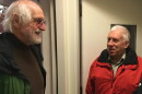 Man in a green coat and brown sweater standing in a doorway speaking to a man in a red coat in front of an interior door.