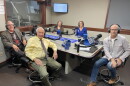 Five smiling people wearing headphones sit around a table in a radio talk studio: a man front left has short grey hair and is wearing glasses, a tan collared shirt under a grey and red argyle sweater and grey suit coat, black pants, and brown shoes; a man front center has short white hair and is wearing a yellow button-down shirt, a grey patterned tie, green pants, and brown shoes; a woman back left has short blonde hair and is wearing glasses and a brown top; a woman back center has curly light brown hair and is wearing glasses and a bright blue top; a man at right has very short dark hair and is wearing glasses, a white shirt under a beige coat, jeans, and brown shoes