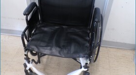 A white plastic bag is tied across the bottom of a black wheelchair at Women's Huron Valley Correctional Facility, the state's only women's prison where the footrests should be. The wheelchair is on a brown tiled floor against a white wall.