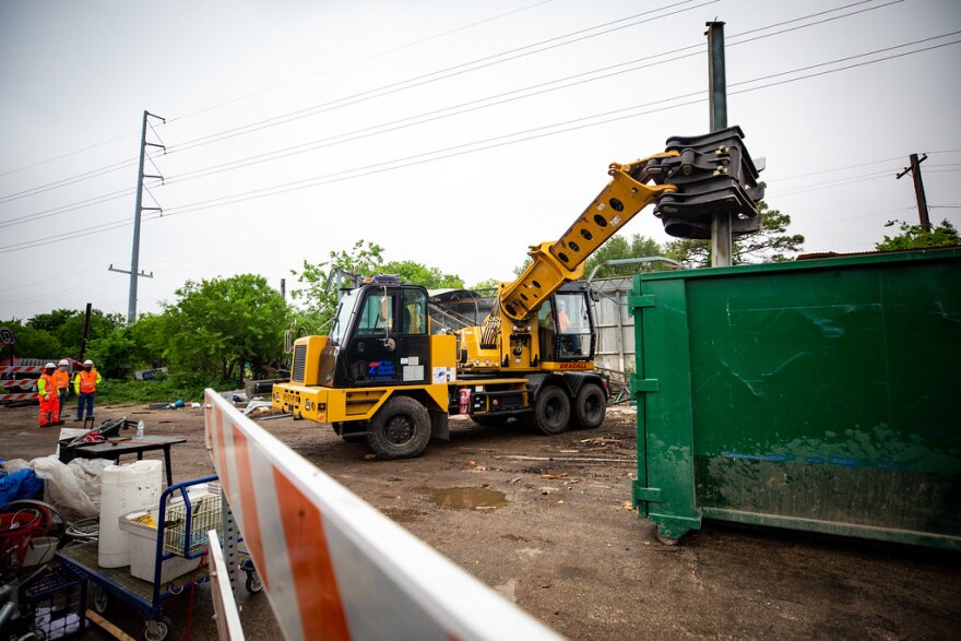 An excavator operated by TxDOT dumps a support beam from a garage into a dumpster. The land on which Esperanza Community sits used to be a maintenance facility for TxDOT until it was repurposed.