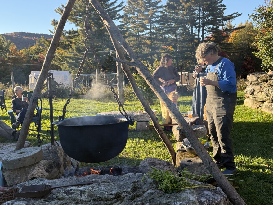 A steaming black cauldron hanging on a rack above a fire with someone standing drinking a beer nearby