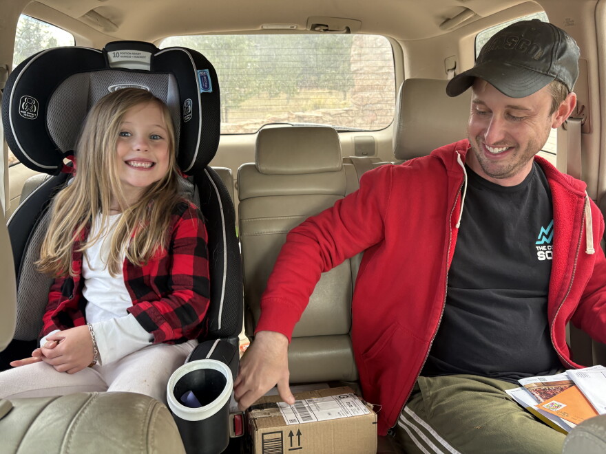 A child, smiling, sits in her car seat, next to her father. 