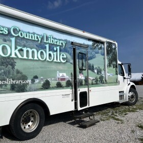 The Holmes County Library Bookmobile is parked in a gravel lot. It's a white truck covered in a photo of rolling hills with a red farm house.