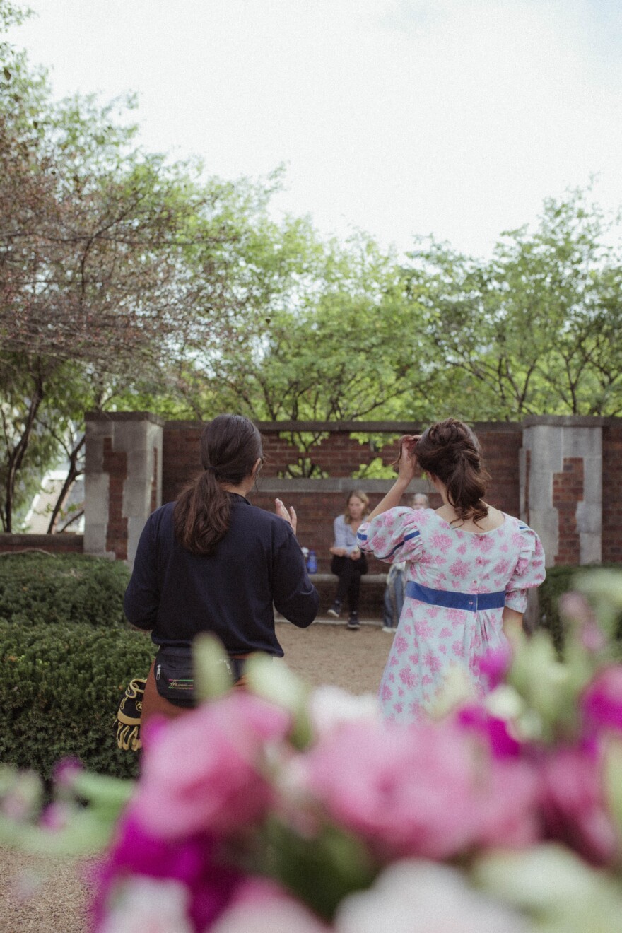 Two women stand in a garden with their backs to the camera on a movie set. 