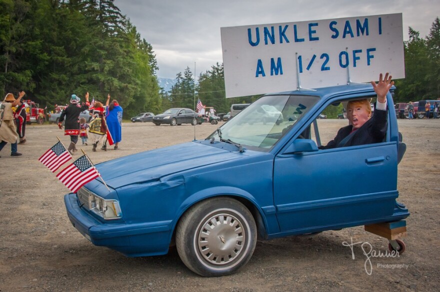 A man in a trump mask holds a sign up through the window of the front half of a blue sedan. The sign says "Uncle Sam I am 1/2 off"
