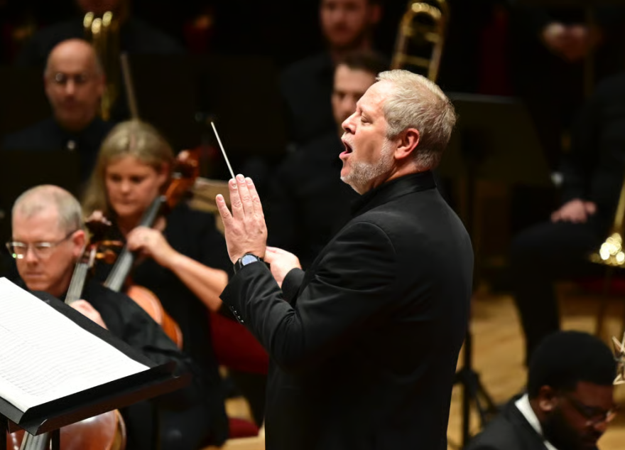 Man in suit with grey hair conducts group of people holding instruments 