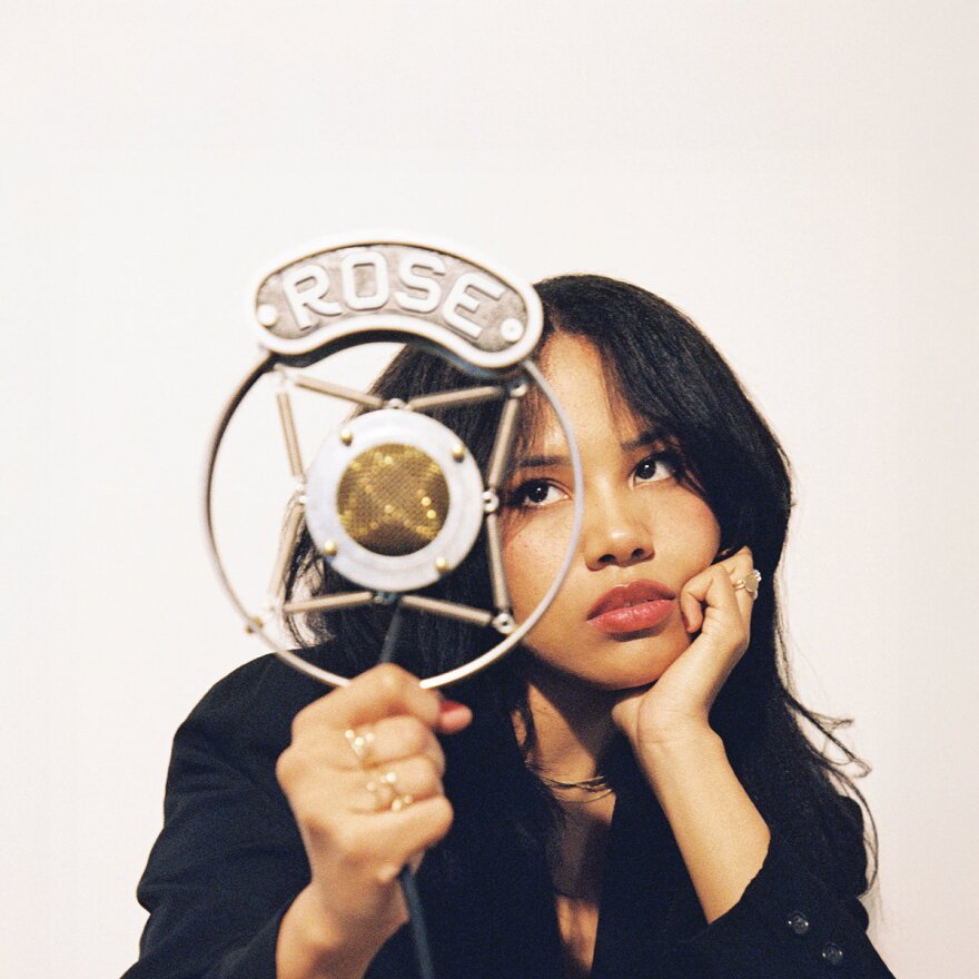 A woman sits behind an old fashioned microphone with the word 'ROSE' above it. She looks bored with her hand resting under her chin while the other hand is holding the microphone.