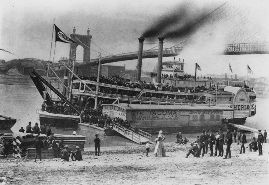 A black and white photograph of a riverboat with a suspension bridge behind it.