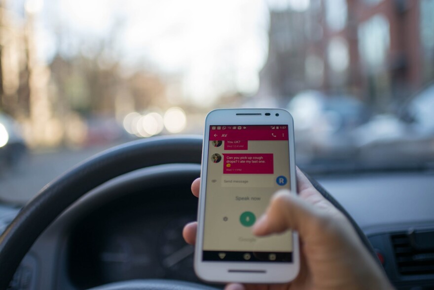 First person perspective of a driver who is texting. The driver holds a phone with a text message displayed on the screen. It reads, "Are you okay?" and "Can you pick up cough drops? I ate my last one." The driver's thumb is poised to start typing.