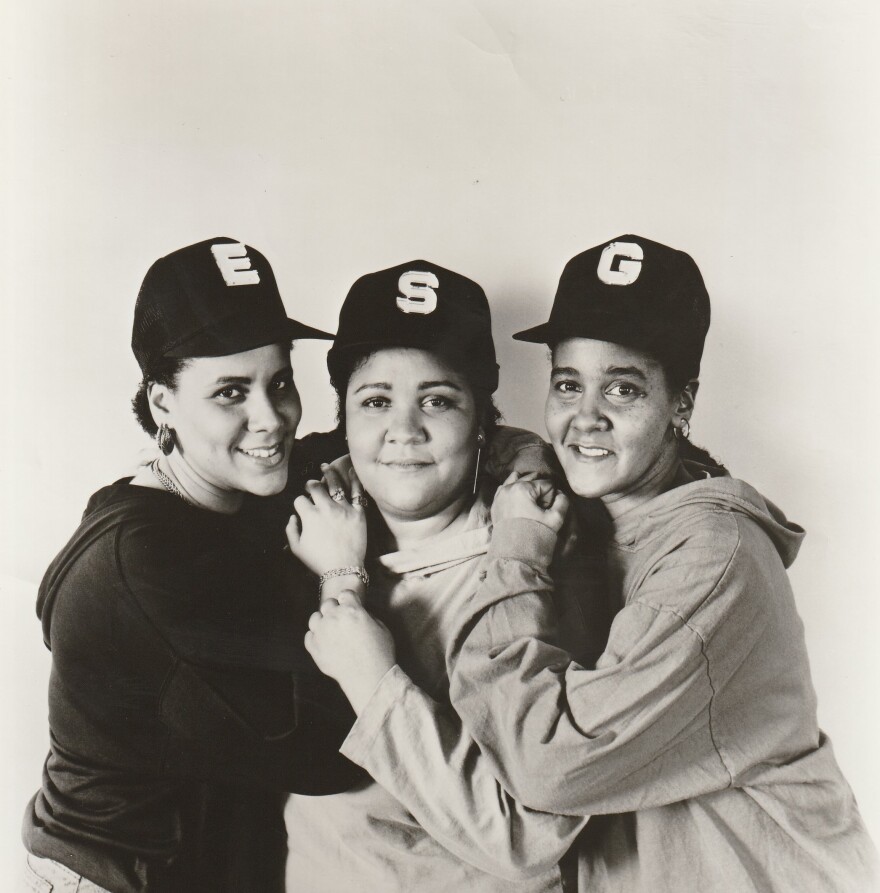The three founding members of ESG pose for the camera with their arms resting on each others' shoulders. They wear black baseball hats: one that says E, one that says S, and one that says G. They also wear hoodies. The photo is staged and in black and white.