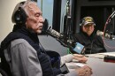 Two men sit inside a radio studio. They are seated at microphones. One on left, Dr. Gene Chavez, is talking while another Frank Hernandez, is listening.