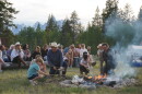Adults and kids kneel and squat around a smoky campfire, roasting marshmallows. People sit in the background, in front of a view of rocky mountains.