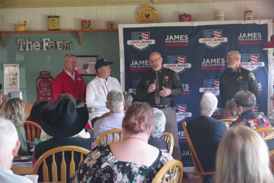 In front of a crowd of people inside a room with farmhouse-themed decor, a man in a sheriff's uniform speaks into a microphone. Another man in a sheriff's uniform stands next to him, and two men in white and red shirts stand off to the side. 