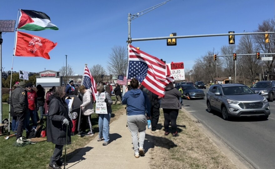 Protesters walk along the sidewalk waving flags -- some are American flags turned upside down, one palestinian flag is stuck in the ground next to the sidewalk. 