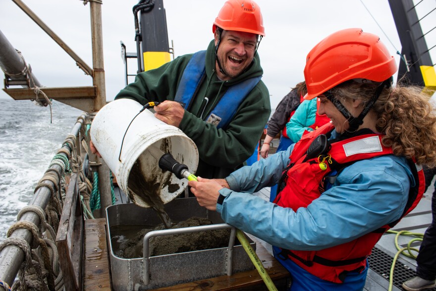 Person empties a bucket into a sieve on a boat while another hoses out the bucket.