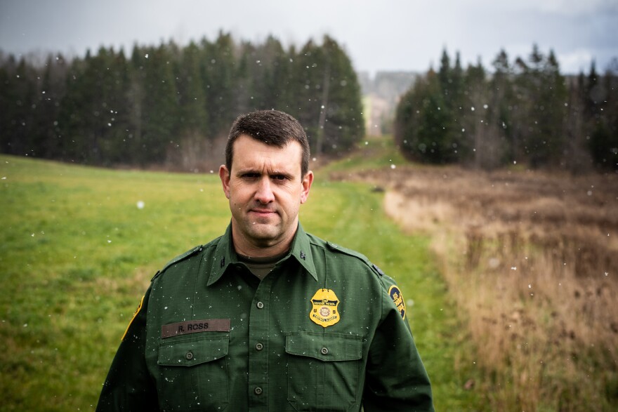 Richard Ross, patrol agent in charge of the United States Border Patrol's Newport Station, stands near the international boundary, marked by the slash in the forest.