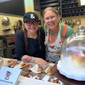 Alicia Dawn, wearing a black shirt and cap, and Erica Idso-Weisz, wearing a colorful apron, smile together behind the counter at Happy Luncheonette in Medford. Small plated appetizers sit in front of them, with a covered pastry stand in the foreground and the restaurant kitchen in the background.