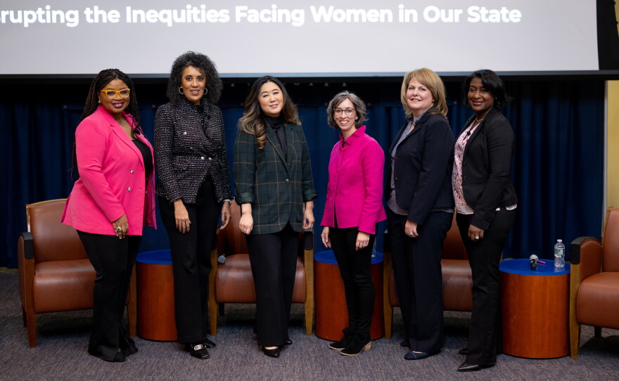 Six women stand in front of a group of chairs and a projector screen. From left to right, Khalilah Brown-Dean wears a pink jacket and black pants, Karen DuBois-Walton wears a dark jacket and black pants, Emily Byrne wears a green jacket and black pants, Michelle Riordan-Nold wears a pink jacket and black pants, Jenny Steadman wears a dark jacket and dark pants and Karen Lott wears a dark jacket and black pants.