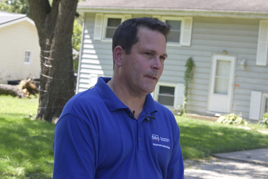 Man in a blue collared shirt standing in front of a two-story home with a front yard and a fallen tree in the background.