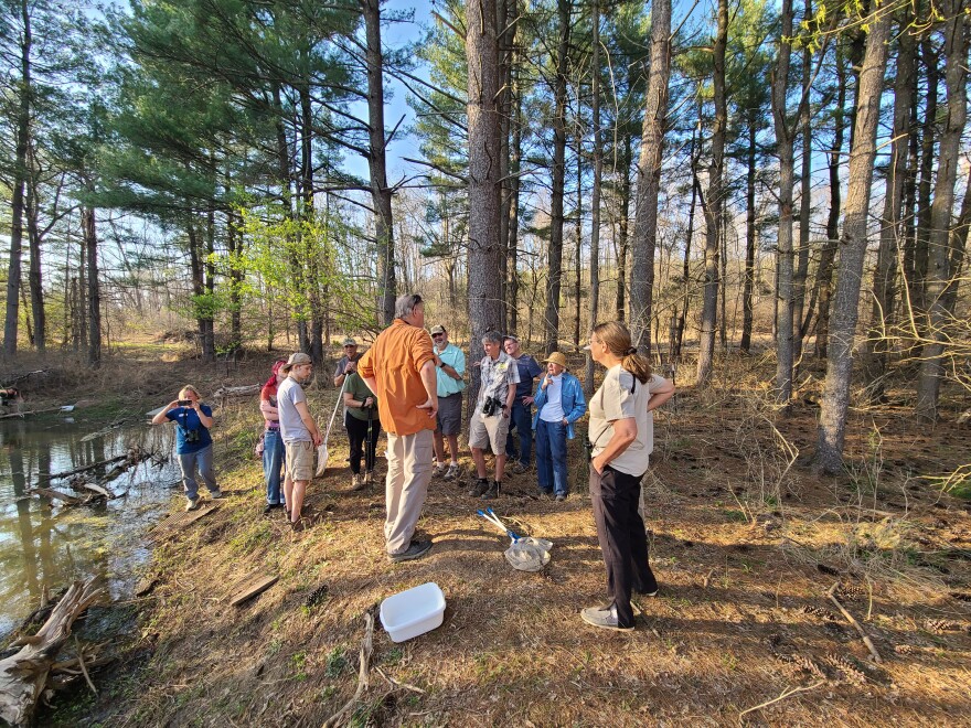 man stands at center of semi-circle of people at the edge of a pond