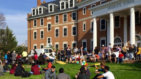 Students sit and stand on a lawn, eating and talking. 